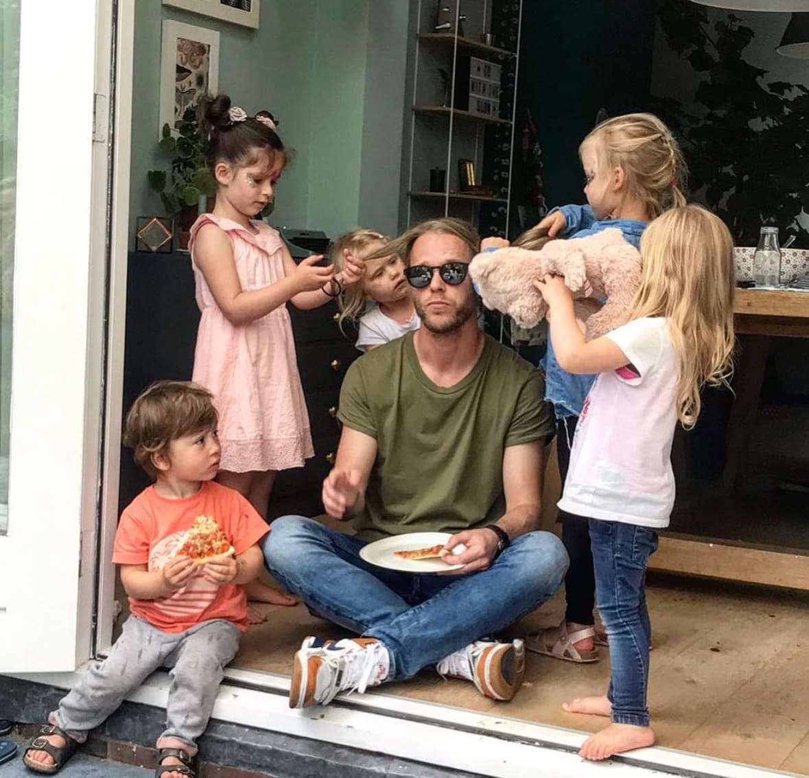 Floris sitting cross-legged with a slice of pizza, completely unbothered while five kids do his hair